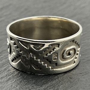 Op de afbeelding: Een zilveren ring met een gegraveerd patroon van geometrische vormen en een spiraalvormig ontwerp. De ring heeft een brede band en een gepolijste afwerking. De ring is waarschijnlijk gemaakt van sterling zilver.