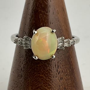 May include: A silver ring with an oval opal gemstone, displaying a pearlescent effect. The ring has small, clear accent stones on either side of the opal. The ring is displayed on a wooden stand. The opal has a warm, creamy colour.