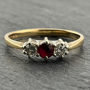Könnte beinhalten: Ein goldener Ring mit drei Edelsteinen. Der Mittelstein ist ein oval geschliffener, tiefroter Rubin, flankiert von zwei runden, klaren Diamanten. Der Ring liegt auf einer dunkelgrauen Oberfläche.