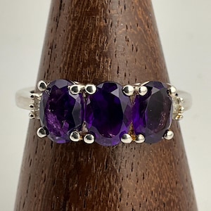 May include: A silver ring with three oval-shaped amethyst gemstones set in a row. The ring is on a wooden display stand.