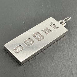 May include: A rectangular silver pendant with a loop at the top. The pendant is stamped with several hallmarks, including a lion passant, an anchor, and the initials "CJC". The pendant is displayed on a dark surface.