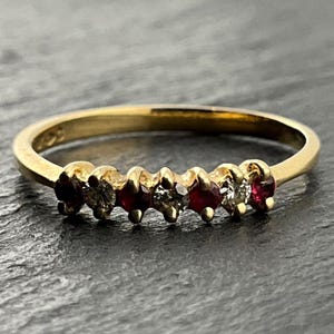 May include: A gold band ring featuring a row of seven alternating round-cut gemstones. The setting includes four deep red rubies and three clear white diamonds arranged in a sequence across the front of the polished yellow gold band.