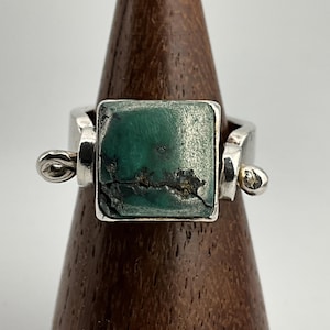 Puede incluir: Anillo cuadrado de plata con una piedra rectangular grande de color verde azulado. La piedra tiene una superficie texturizada con vetas oscuras. El anillo tiene un diseño único con detalles decorativos de plata. El anillo se exhibe en un soporte de madera.
