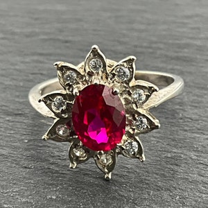 May include: A silver ring with a large, oval-shaped, bright red ruby surrounded by smaller, clear, round stones. The stones are set in a flower-shaped design.