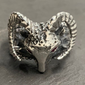 May include: A silver ring with a detailed ram head design. The ram's horns are curled and the eyes are set with red stones.
