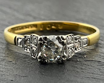 May include: A gold ring featuring a central round-cut diamond set in a white metal head, flanked by smaller diamonds on each side. The band is smooth yellow gold, and the setting includes intricate metalwork details surrounding the stones.