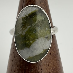 Peut inclure: Bague argentée ornée d'une pierre ovale verte et blanche. La pierre présente un aspect marbré. La bague est présentée sur un support en bois. Le design est simple et élégant.