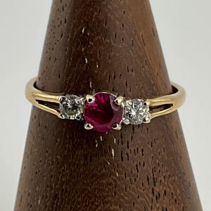 May include: A gold ring featuring a central, round, deep red gemstone, with two smaller, clear gemstones either side. The ring's band splits on either side of the central stone. The ring is displayed on a wooden stand.