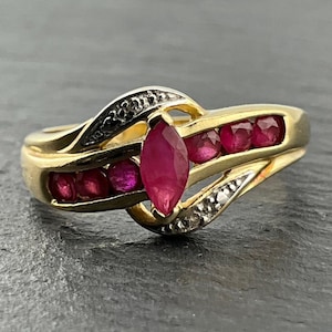 May include: A gold ring featuring a central marquise-cut red ruby flanked by smaller round-cut red rubies set within a bypass-style band. The band includes two curved sections accented with small, clear, sparkling stones along the outer edges.