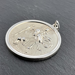 Puede incluir: Colgante redondo plateado con un relieve detallado de un santo que lleva a un niño a través del agua. El colgante tiene un borde en relieve y un lazo para una cadena. El diseño es intrincado y el metal tiene un acabado pulido.