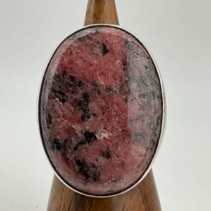 May include: A large oval ring featuring a pink gemstone with black inclusions, set in a silver-colored metal band. The gemstone has a polished surface and is mounted on a wooden display stand.