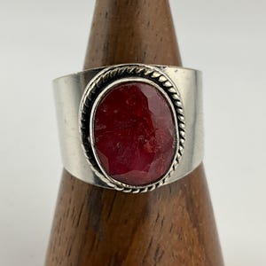 May include: A wide silver ring featuring a large, oval-cut, deep red gemstone. The gemstone is set within a twisted rope-like border. The ring is displayed on a wooden ring holder against a white background.