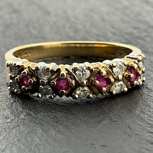 May include: A yellow gold band ring featuring a repeating pattern of four round red rubies alternating with clusters of small clear white diamonds set in a cross-over design. The metal band has a polished finish and visible hallmarks on the inner side.