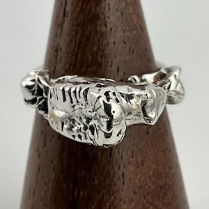 以下が含まれることがあります： 生き物の顎や歯を描いた可能性のある、テクスチャ加工が施されたデザインのシルバーリング。リングは、金属の複雑な質感と形状に焦点を当てた、詳細な3次元の外観をしています。リングは木製のスタンドに展示されています。