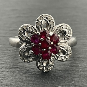 Puede incluir: Un anillo de plata con un diseño de flor que presenta un grupo de rubíes rojos en el centro y piedras blancas más pequeñas alrededor de los bordes.