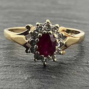 May include: A gold ring featuring a central oval-cut deep red ruby surrounded by a halo of small, clear, round-cut stones. The band is polished gold with a split-shank design that connects to the ornate, prong-set cluster setting at the center.