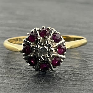 Puede incluir: Un anillo de oro con un engaste en racimo que presenta una piedra central clara rodeada por ocho rubíes de color rojo intenso. El racimo está montado sobre una banda de oro pulido. El diseño crea una disposición floral con los rubíes en halo.