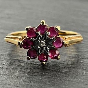 May include: A gold ring featuring a cluster of round, deep pink rubies arranged in a floral pattern around a central dark, star-shaped setting with a single small, clear, round-cut diamond at its very centre, set on a polished gold band.