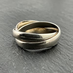 Könnte beinhalten: Ein silberner Ring mit drei ineinander verschlungenen Bändern. Der Ring besteht aus drei Bändern, die ineinander verschlungen sind und zu einem glänzenden Finish poliert wurden.
