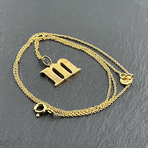 May include: A gold-coloured necklace featuring a pendant in the shape of the letter "m". The pendant is a capital letter with a slightly aged appearance. The chain is delicate and has a clasp closure. The necklace is displayed on a dark surface.