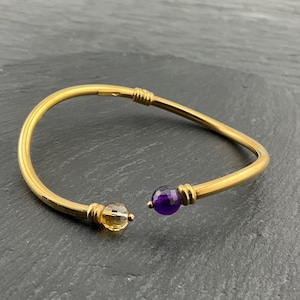 May include: A gold-colored open cuff bracelet with a citrine and an amethyst gemstone. The bracelet has a curved design with a clasp closure. The gemstones are round and faceted.