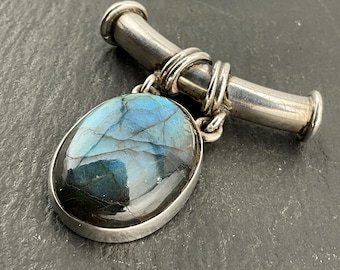 Puede incluir: Broche plateado con un colgante de piedra labradorita ovalada. La piedra muestra tonos iridiscentes azules y verdes. El broche tiene un diseño de barra y lazo, con la piedra suspendida de la barra.