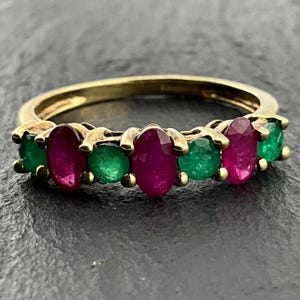 May include: A gold ring featuring an alternating row of three oval-cut red rubies and four round-cut green emeralds set in a prong setting. The band is crafted from polished yellow gold, showcasing a vibrant contrast between the deep red and green gemstones.