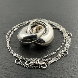 May include: A silver pendant necklace with an intertwined, rounded design. The pendant hangs from a delicate chain with a lobster clasp. The necklace is presented on a dark, textured surface.