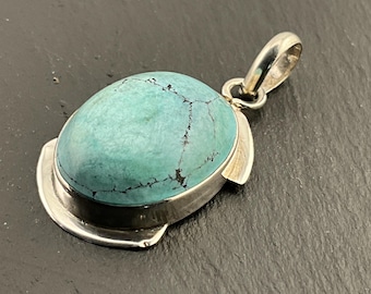 May include: Oval turquoise pendant with a silver bail. The turquoise stone has dark veining and is set in a silver bezel. The pendant is a classic jewelry piece.