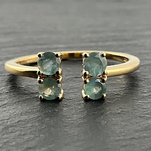 May include: A gold open-style ring featuring four round-cut, teal-coloured gemstones. The stones are arranged in two pairs, with two gems set vertically on each side of the open gap in the band, held in place by small gold prongs.