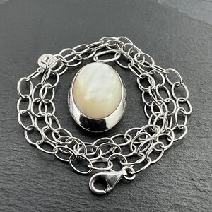 May include: A silver-toned necklace with an oval pendant featuring a pearlescent, off-white stone. The pendant is set in a polished silver bezel. The chain is composed of interconnected oval links and includes a lobster clasp.