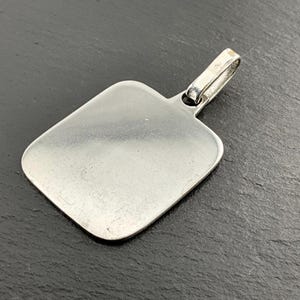 Peut inclure: Un pendentif argenté de forme rectangulaire aux coins arrondis. Il est doté d'une petite boucle en haut pour une chaîne. La surface du pendentif est lisse et réfléchissante.