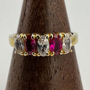 以下が含まれることがあります： 5つの楕円形の宝石をあしらったゴールドリング。透明な石と鮮やかな赤い石が交互に並び、ゴールドのバンドにセットされています。宝石は小さなゴールドの爪でしっかりと固定されています。