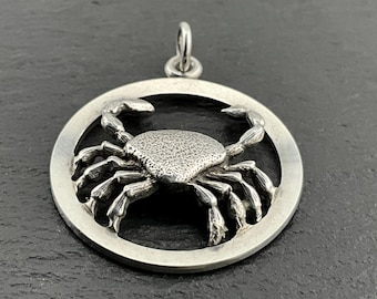 May include: A silver pendant featuring a detailed crab design within a circular frame. The crab is textured and has a three-dimensional appearance.