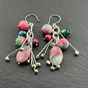 Könnte beinhalten: Ohrringe mit Anhängern mit einer Ansammlung von rosa, grünen und lila Perlen. Die Ohrringe haben ein silberfarbenes Metalldesign mit Hakenverschluss. Die Perlen sind in verschiedenen Formen und Größen gehalten und schaffen ein einzigartiges Accessoire.