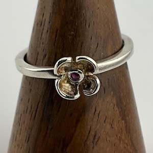 Könnte beinhalten: Ein silberner Ring mit einem Blumendesign und einem kleinen rosafarbenen Edelstein in der Mitte.