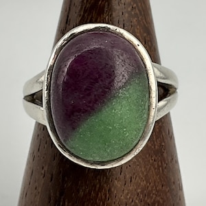 May include: A silver ring featuring an oval-shaped stone divided into two sections: a deep purple and a light green. The ring has a split band design, adding a unique touch to the jewelry.