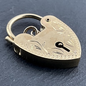 May include: A gold-toned heart-shaped padlock charm featuring an engraved floral pattern on the front surface. The charm includes a curved shackle at the top and a small jump ring attached to the back, with a keyhole-shaped cutout in the center.