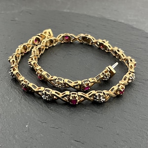 Puede incluir: Una pulsera de oro con un diseño de eslabones entrelazados en forma de X, acentuada con rubíes rojos redondos y pequeños diamantes transparentes alternados, engastados en monturas de metal plateado a lo largo de la cadena circular.
