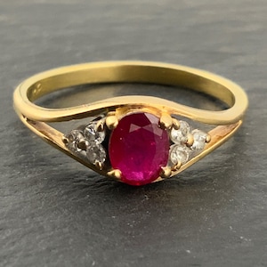 Może przedstawiać: Złoty pierścionek z owalnym rubinem w centrum, otoczonym małymi, błyszczącymi kamieniami. Pierścionek ma delikatny, płynny design z polerowaną złotą obrączką. Rubin ma głęboki, bogaty czerwony kolor.