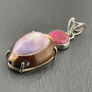 Può includere: Un ciondolo d'argento con un cammeo di conchiglia marrone e viola e una pietra preziosa rossa. Il ciondolo è su uno sfondo nero.
