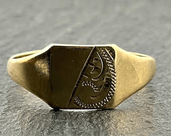 May include: A gold signet ring with a rectangular face and a detailed design. The ring has a polished finish and a classic, vintage style. The face of the ring features an engraved pattern.
