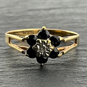 May include: A gold ring featuring a floral-shaped cluster setting. The centre of the flower contains a small, clear, faceted stone surrounded by six round, dark black stones. The band is made of polished yellow gold with a split-shank design.