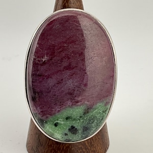 Peut inclure: Une bague ovale avec une pierre précieuse en rubis zoisite polie. La pierre présente une teinte bordeaux profond avec des inclusions vertes et noires, sertie dans une lunette argentée.