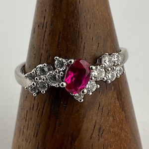 May include: A silver ring featuring a vibrant oval-cut ruby center stone, flanked by clusters of smaller, round, clear gemstones. The ring's band has a delicate, flowing design, creating a floral-like appearance. The gemstones sparkle against the silver setting.