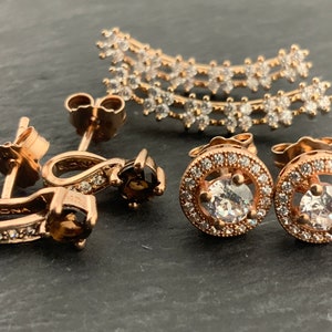 May include: A set of three pairs of rose gold earrings with clear and brown stones. The earrings are all different styles, with one pair featuring a curved design, another pair featuring a round design, and the third pair featuring a teardrop design.