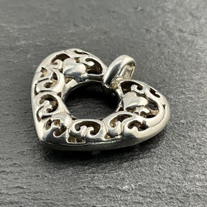 May include: A silver heart-shaped pendant with an openwork design. The pendant has a loop at the top for a chain. The heart is decorated with swirling patterns and small heart shapes. The pendant is displayed on a dark grey surface.