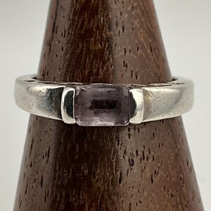 May include: A silver ring featuring a rectangular, pale purple gemstone. The band is thick and has a simple, modern design. The gemstone is set in a bezel setting. The ring is displayed on a wooden stand.