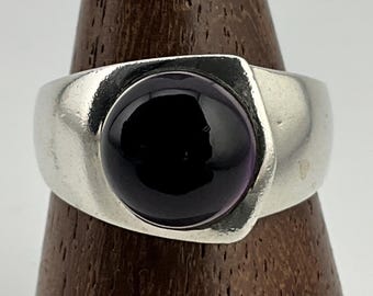 May include: A silver ring with a large, round, dark purple gemstone. The ring has a wide band and a bezel setting that holds the stone securely. The gemstone is smooth and polished, reflecting light.