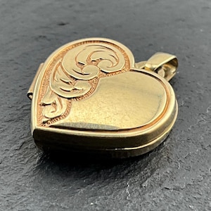 Könnte beinhalten: Ein goldfarbenes, herzförmiges Medaillon mit einem eingravierten Schnörkelmuster auf der linken Seite und einer glatten, polierten Oberfläche auf der rechten Seite. Das Medaillon hat oben eine kleine Metallöse zur Befestigung an einer Kette.